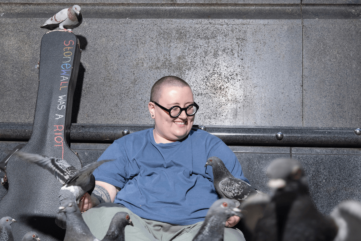 Maddie Morris, sat with their guitar flight case that says 'Stonewall was a riot', they are smiling while feeding pidgeons.